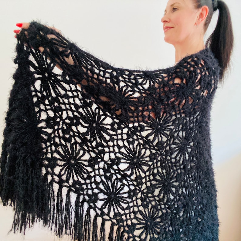 May include: A black, open-weave shawl with a floral pattern and fringe. The shawl is made of a soft, fuzzy material. The floral design is repeated throughout the shawl. The fringe hangs along the bottom edge of the shawl.