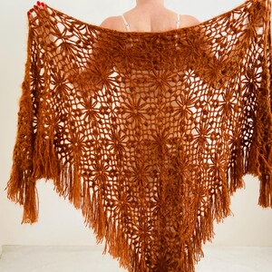 May include: A handmade, open-weave shawl in a warm, reddish-brown hue. The shawl features a floral pattern throughout, with a fringed edge. The intricate design and color make it a unique accessory.