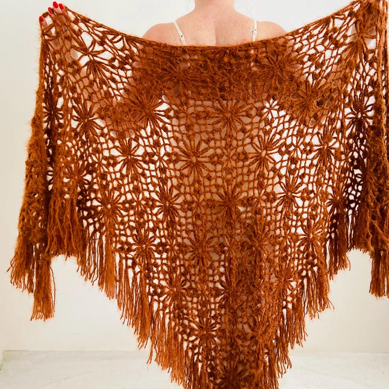 May include: A handmade, open-weave shawl in a warm, reddish-brown hue. The shawl features a floral pattern throughout, with a fringed edge. The intricate design and color make it a unique accessory.
