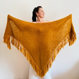 Puede incluir: Un chal de punto amarillo mostaza con flecos. El chal está drapeado sobre los hombros de una persona.