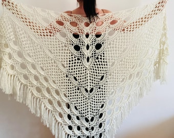 May include: Cream-colored, open-weave crochet shawl with a triangular shape. The shawl features a central, detailed pattern and a fringe along the edges. The design includes large circular openings and a decorative pattern.
