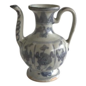 Op de afbeelding: Antieke keramische kan in wit en blauw, met een gebogen tuit en handvat. Het heeft bloempatronen en een voetstuk. Het ontwerp is een klassiek voorbeeld van vintage aardewerk.