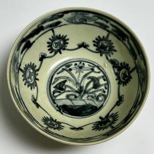 Puede incluir: Cuenco de cerámica antigua con interior color crema y diseño floral azul oscuro. El cuenco presenta un motivo floral central y un patrón repetido de flores estilizadas alrededor del borde. El diseño está pintado a mano, mostrando la artesanía de la época.