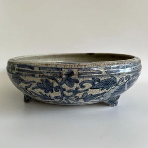 Könnte beinhalten: Eine niedrige, runde Keramikschale mit blau-weißem Blumendesign. Die Schale hat drei kleine Füße und eine Craquelé-Glasur. Das blaue Blumenmuster umfasst Blätter und Ranken. Die Schale ist wahrscheinlich Vintage oder antik.