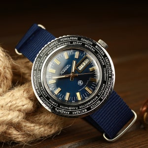 Może przedstawiać: Niebieski i srebrny zegarek na rękę z tarczą zegara światowego. Tarcza zegarka ma niebieski kolor z złotymi akcentami i okienkiem daty. Pasek zegarka to ciemnoniebieski pasek nylonowy.