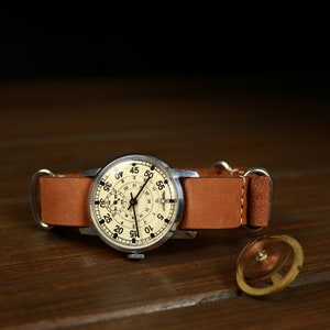 Puede incluir: Un reloj de pulsera plateado con una esfera blanca y marcas negras. El reloj tiene una correa de cuero marrón y está sobre una superficie de madera. Un muelle de reloj está junto al reloj.