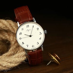 May include: A silver wristwatch with a white face and black hands. The watch has a brown leather band and the face has black Arabic numerals. The watch face also has the word "Mыr" written in cursive.