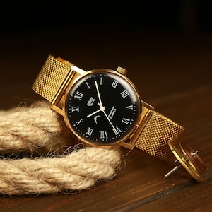 May include: A gold-tone wristwatch with a black face and Roman numeral hour markers. The watch has a gold-tone metal band and a crescent moon design on the face. The watch is on a wooden surface with a piece of rope.