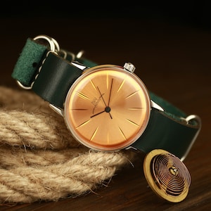 May include: A vintage wristwatch with a round, orange face and silver hands. The watch has a green leather band and is sitting on a brown surface.