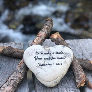 May include: A heart-shaped stone with the inscription "Let's make a trade: Your rock for mine? September 1, 2019" and a gold ring on top, resting on a wooden surface.