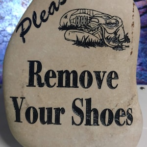 May include: A light brown stone with the words "Please Remove Your Shoes" written in black lettering. A black and white illustration of a shoe is above the text.