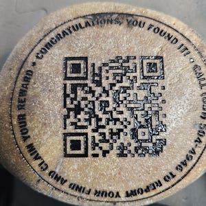 May include: A circular tan stone featuring a black laser-engraved QR code in the center. The text surrounding the QR code reads: CONGRATULATIONS, YOU FOUND IT! CALL (859) 504-4946 TO REPORT YOUR FIND AND CLAIM YOUR REWARD. A & W Engraving is printed below.