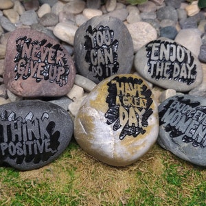 Puede incluir: Seis piedras pintadas con mensajes inspiradores. Las piedras son grises, marrones y doradas con letras negras. Los mensajes son "Think Positive", "Never Give Up", "You Can Do It", "Have A Great Day", "Enjoy The Day" y "Enjoy Every Moment".