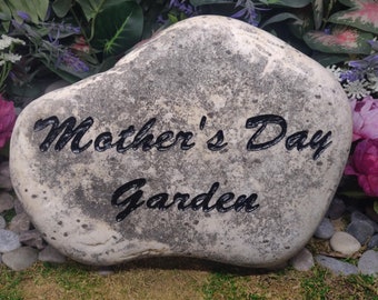 Può includere: Una pietra di fiume liscia di colore grigio chiaro con una consistenza screziata e usurata. Le parole Mother's Day Garden sono incise in un carattere corsivo nero al centro della pietra, creando un contrasto con la superficie.