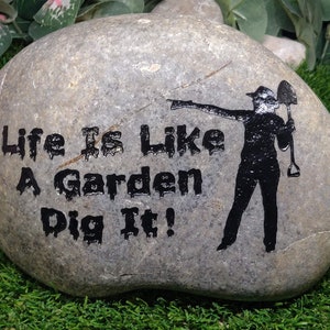 May include: A gray stone with the text "Life is like a garden Dig it!" and a black silhouette of a person holding a shovel.