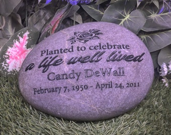 May include: A smooth, oval-shaped gray memorial stone featuring an engraved rose graphic and the text: Planted to celebrate a life well lived, Candy DeWall, February 7, 1950 - April 24, 2011. The lettering and rose are etched in black.
