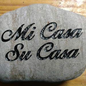 Op de afbeelding: Een grijze steen met de woorden "Mi Casa Su Casa" gegraveerd in zwarte letters.