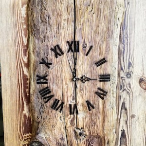 May include: A rustic wooden wall clock with a natural, unfinished appearance. The clock face features black Roman numerals and hands. The clock is made from two pieces of wood joined together, showcasing the wood grain.