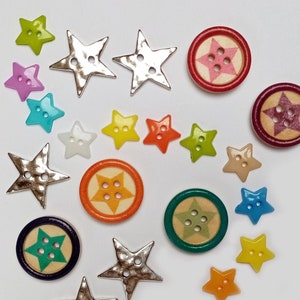 Op de afbeelding: Assortiment van kleurrijke stervormige knopen met verschillende afwerkingen, waaronder zilver, goud en plastic. Sommige knopen hebben een reliëfsterontwerp, terwijl andere een vlak oppervlak hebben.