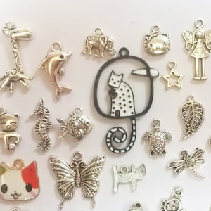 Op de afbeelding: Een verzameling zilverkleurige metalen bedels in diverse vormen, waaronder dieren zoals een giraffe, dolfijn, kat en vlinder, naast bloemen- en geometrische ontwerpen. De bedels zijn gerangschikt op een wit oppervlak.