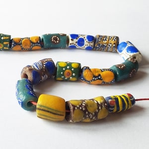 Op de afbeelding: Een streng kleurrijke, handgemaakte kralen. De kralen hebben een mix van vormen en maten, met gele, blauwe, groene en witte tinten en ingewikkelde patronen. De kralen zijn geregen op een dun rood koord, wat een uniek accessoire creëert.