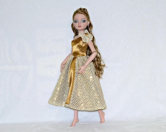 May include: A doll wearing a gold sequined dress with a one-shoulder bodice and a matching sash. The doll has long, light brown hair and is adorned with a decorative flower on the shoulder. The dress sparkles with gold sequins.