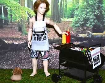May include: A doll wearing a white apron with the text "GO TOPLESS" and a black Jeep graphic, paired with patterned shorts. A black grill with condiments and a towel that says "BORN TO GRILL" is nearby. The scene is set outdoors.