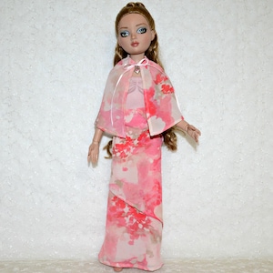May include: A doll wearing a pink floral print dress and matching cape. The dress is floor-length with a layered design, and the cape is sheer with a bow at the neck. The doll has blonde hair and blue eyes, and is a fashion doll.