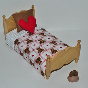 May include: A miniature wooden bed with a brown pillow and a red heart-shaped pillow. The bedspread is white with a pattern of brown teddy bears and red hearts. A small, brown, rounded object sits near the bed.