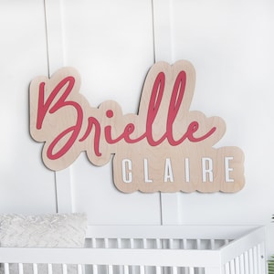 May include: A white crib with a white and gray patterned sheet. A wooden name sign with the name "Brielle Claire" in red script is on the wall behind the crib.
