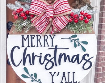 May include: A round wooden Christmas sign with the words "MERRY Christmas Y'ALL" in black script. The sign is decorated with a red and white striped bow, faux berries, and greenery. The top portion is brown wood.