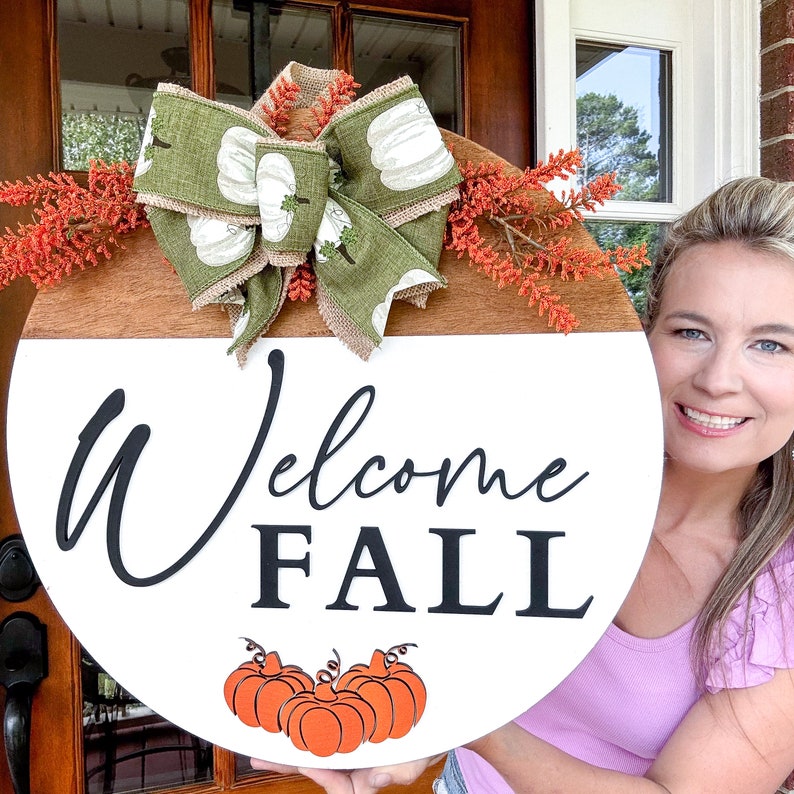 May include: Welcome Fall round wooden sign with a white circle in the center. The words 'Welcome Fall' are written in black. There are three orange pumpkins at the bottom. A green and white bow with burlap is at the top.  Fall-themed decor.