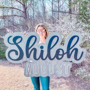 May include: A wooden sign with the name "Shiloh" in blue cursive lettering and the month "August" in white lettering on a gray background.