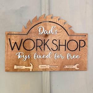 May include: A wooden sign with the text "Dad's Workshop Toys fixed for free" in white lettering. The sign is shaped like a saw blade and has a brown wood grain finish. There are three smaller wooden pieces attached to the bottom of the sign, each with a different tool and a name engraved on them: "Bridget", "Bronx", and "Barnett".