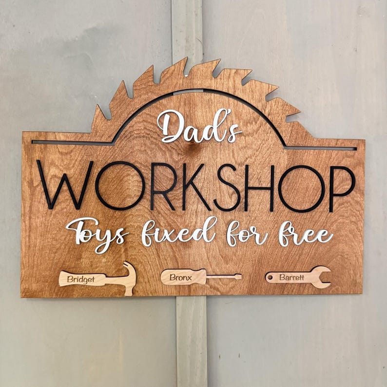 May include: A wooden sign with the text "Dad's Workshop Toys fixed for free" in white lettering. The sign is shaped like a saw blade and has a brown wood grain finish. There are three smaller wooden pieces attached to the bottom of the sign, each with a different tool and a name engraved on them: "Bridget", "Bronx", and "Barnett".