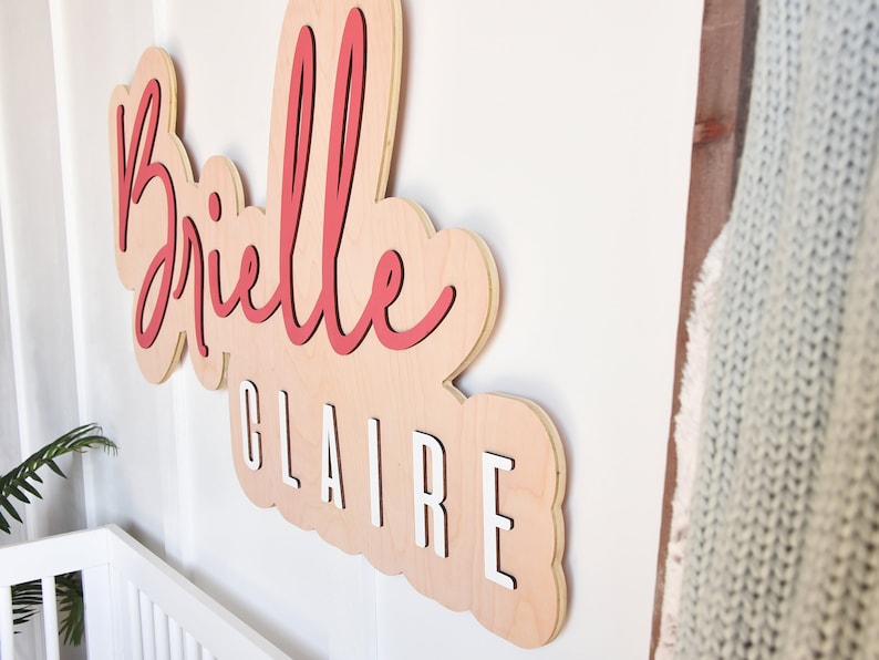 May include: A wooden sign with the names "Brielle Claire" in a cursive font. The name "Brielle" is in red and the name "Claire" is in white.