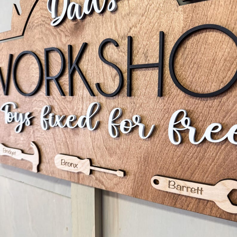 May include: A wooden sign with the text "Dad's Workshop Toys fixed for free" in white lettering. The sign has a black outline and features three wooden tools with the names "Bridget", "Bronx", and "Barrett" engraved on them.