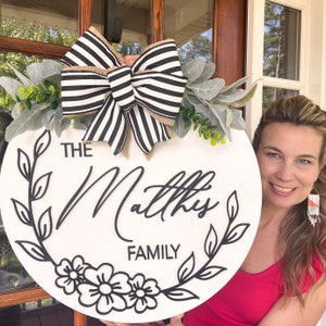 Puede incluir: Un letrero redondo de madera blanca con un lazo a rayas blanco y negro, hojas verdes y detalles florales negros. El letrero dice "The Mathis Family" en letras cursivas negras.