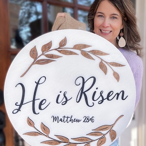May include: A circular, white wooden sign featuring the text "He is Risen" in black script, and "Matthew 28:6" below. The sign is decorated with brown wooden leaf accents. The sign is being held up by a person.