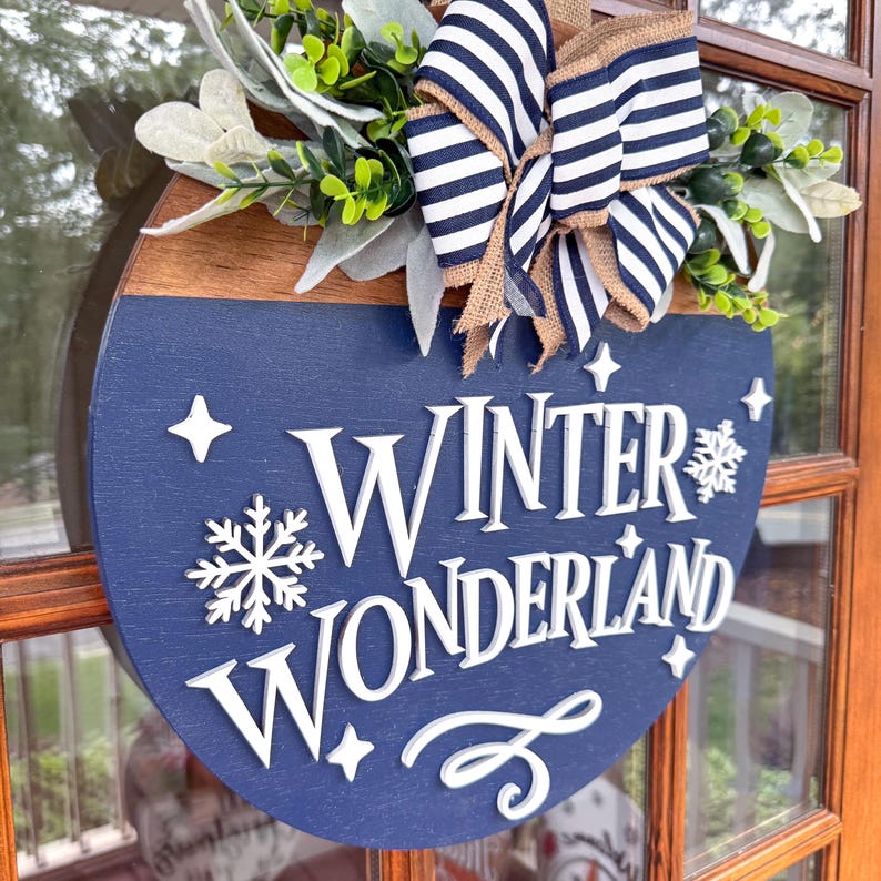 May include: A round wooden sign with a navy blue base and the words "Winter Wonderland" in white. The sign is decorated with a bow with navy and white stripes, greenery, and a snowflake.