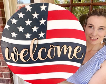May include: A wooden welcome sign painted with an American flag design. The sign has red and white stripes and blue stars. The word "welcome" is written in a white script font across the center of the sign.