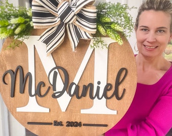 Puede incluir: Un colgador de puerta redondo de madera con una letra M blanca y el nombre "McDaniel" en escritura negra. El colgador está decorado con un lazo a rayas blanco y negro y vegetación. El texto "Est. 2024" está escrito debajo del nombre.