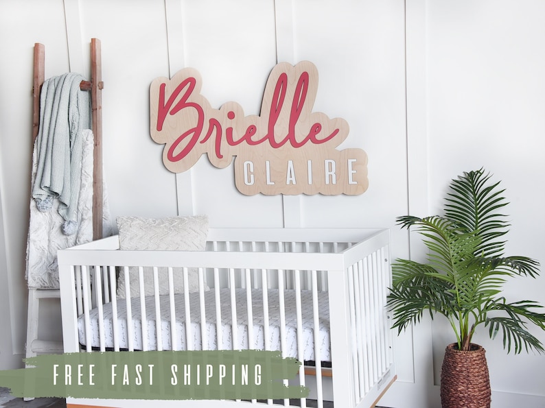 May include: A white crib with a white and gray patterned sheet. A wooden name sign with the name "Brielle Claire" in red script is on the wall behind the crib.