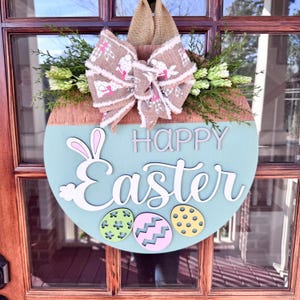 Peut inclure: Une suspension de porte ronde en bois avec un fond bleu et brun. Les mots "Joyeuses P&acirc;ques" sont &eacute;crits en blanc avec une silhouette de lapin. La suspension est d&eacute;cor&eacute;e d'un n&oelig;ud en toile de jute brun et d'accents floraux verts.