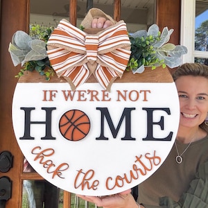 Op de afbeelding: Een ronde houten deurhanger met een witte achtergrond en een oranje-witte gestreepte strik. De tekst "Als we niet thuis zijn, check de courts" staat in zwarte en oranje letters met een basketbalafbeelding in het midden.