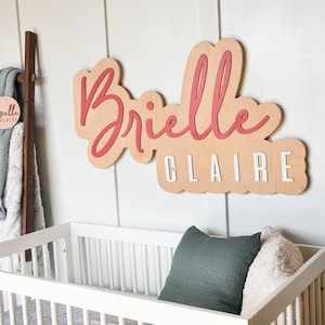 May include: A wooden sign with the name "Brielle Claire" written in red and white paint. The sign is hanging on a white wall above a white crib with a green and white pillow.