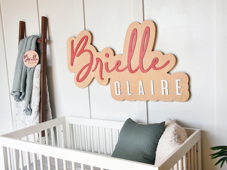 May include: A wooden sign with the name "Brielle Claire" written in red and white paint. The sign is hanging on a white wall above a white crib with a green and white pillow.