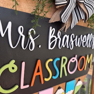 Op de afbeelding: Een zwart schoolbord met de tekst "Mrs. Braswell's Classroom" in witte reli&euml;fletters. De letters in "Classroom" zijn veelkleurig en in reli&euml;f. Het bord is versierd met een groene bladkrans en een zwart-wit gestreepte strik.