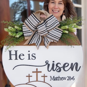 May include: A round wooden Easter sign with the text "He is risen" and "Matthew 28:6". The sign features three wooden crosses and is decorated with a black and white striped bow and greenery.