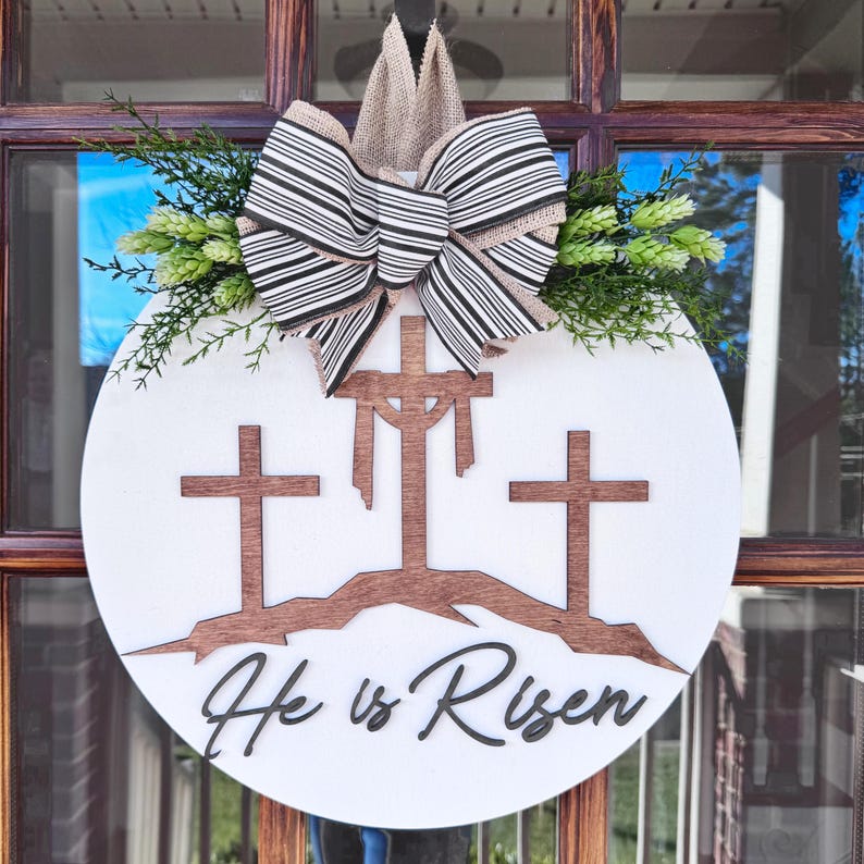 May include: A white round wooden door hanger with three brown wooden crosses and the words 'He is Risen' in black cursive script. The crosses are on a brown hill and there is a burlap and black striped bow with green foliage on top.